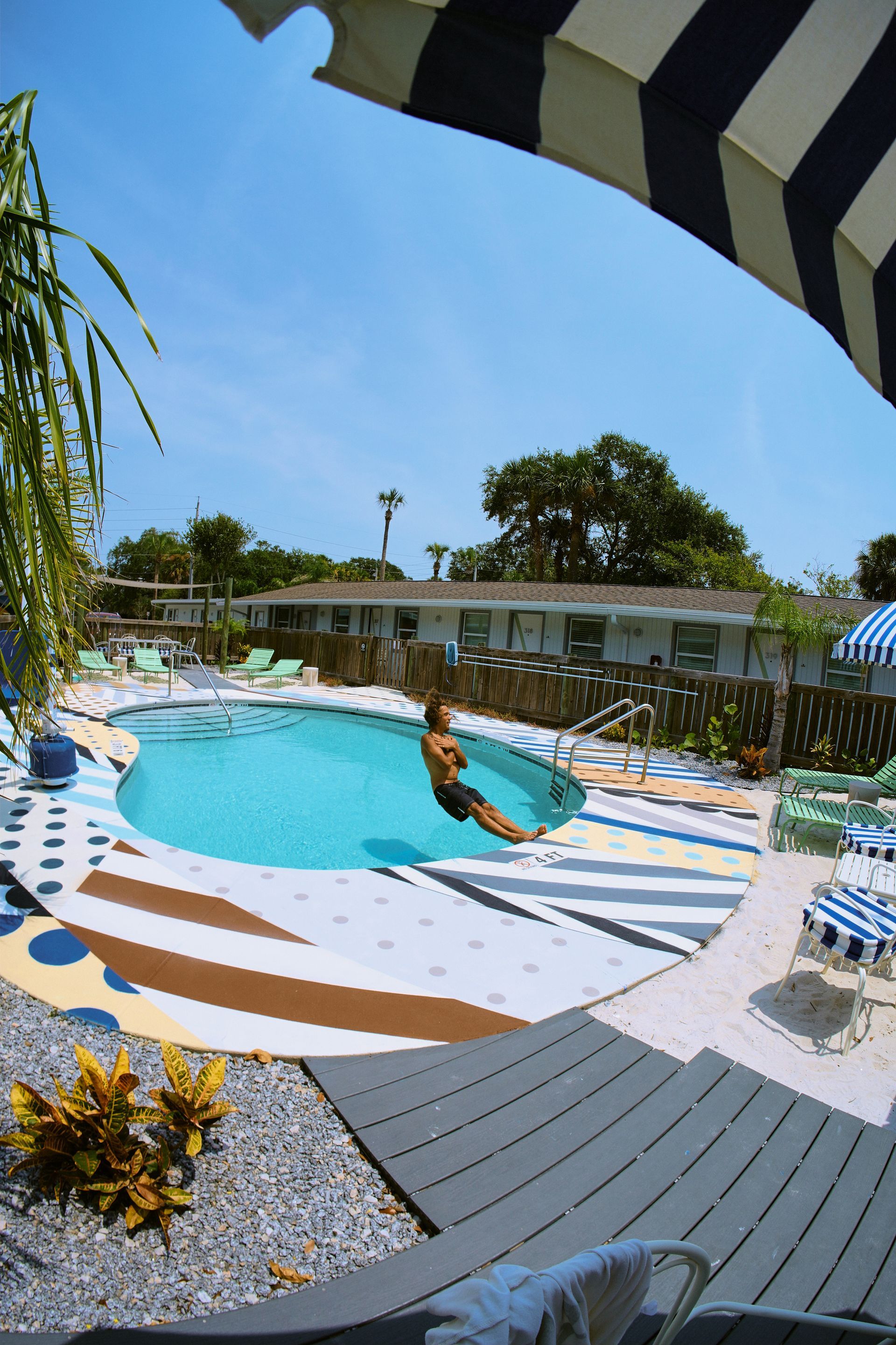 Curved pool with a person floating, striped lounge chairs, and a striped awning in a sunny resort setting