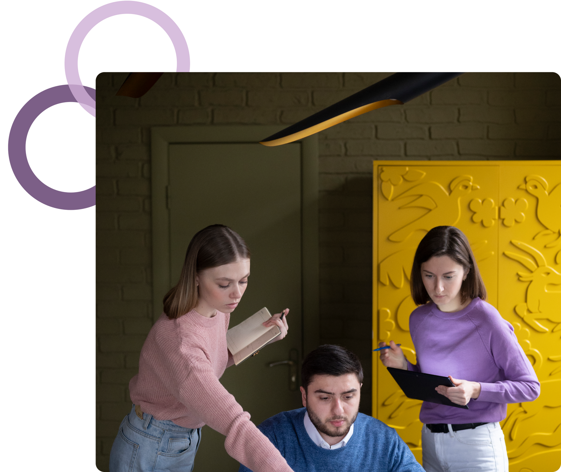 Three people collaborate in a dim room, with one seated and two standing beside a bright yellow wall panel.