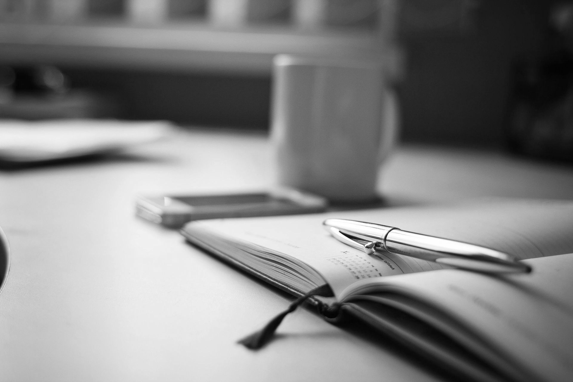Black-and-white desk scene with an open notebook, pen, and coffee mug in the background