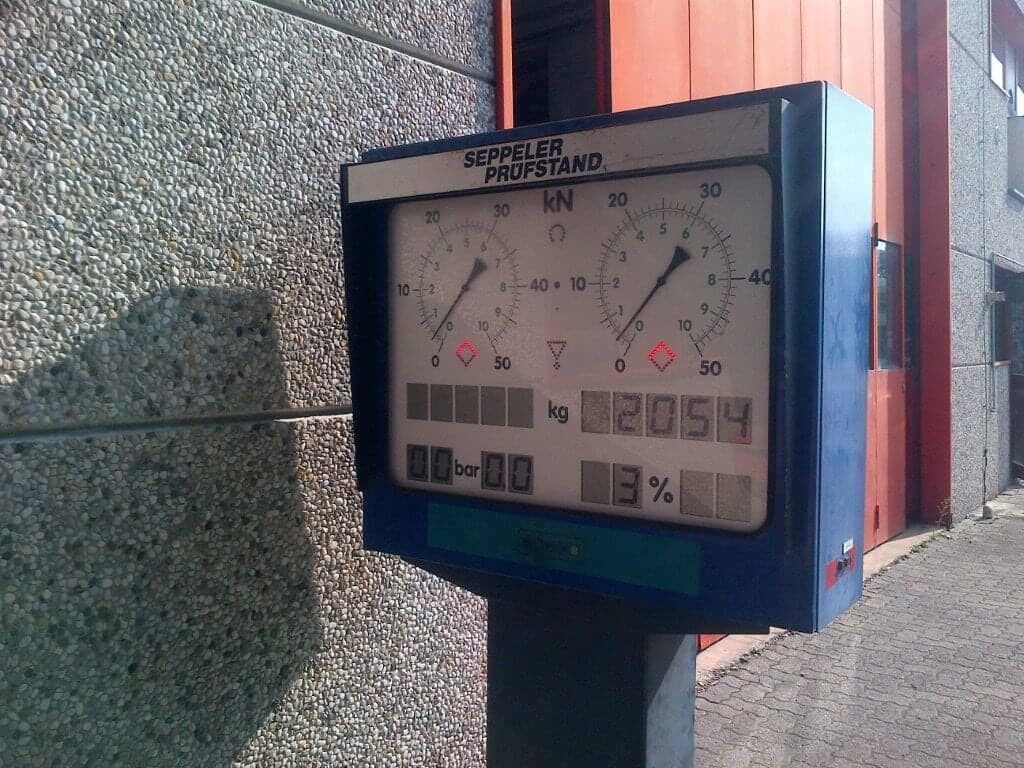 Bilancia industriale blu e bianca su un palo di metallo, con indicatori e display digitale contro una parete strutturata.