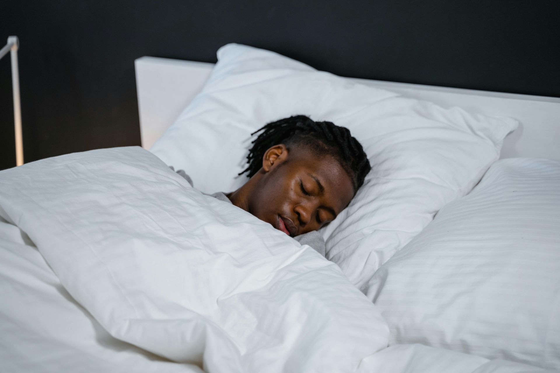 A young Black man asleep in a bed with white sheets and pillows. His eyes are closed, and he is covered by a duvet.