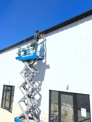 Worker on a blue scissor lift painting a white building wall under a clear blue sky.