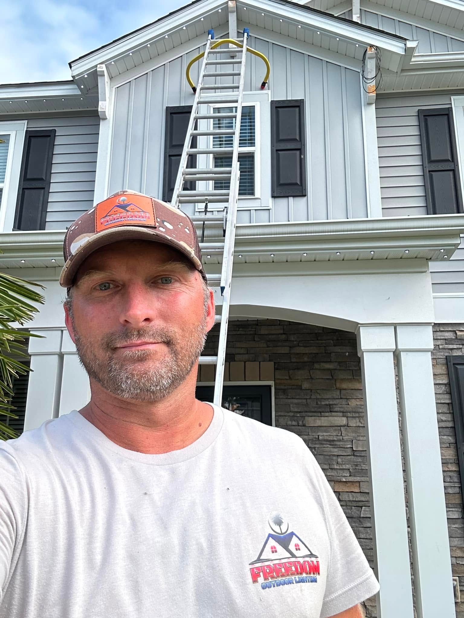Man in cap stands in front of a house with a ladder leaning to the roof.