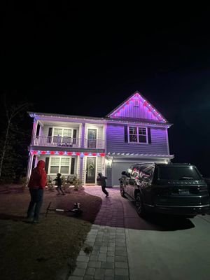 Two-story house lit purple at night with people outside and a car in the driveway