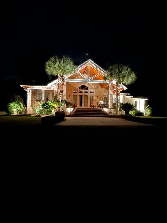 Illuminated house at night with white exterior, orange-lit gable, and trees in the front yard