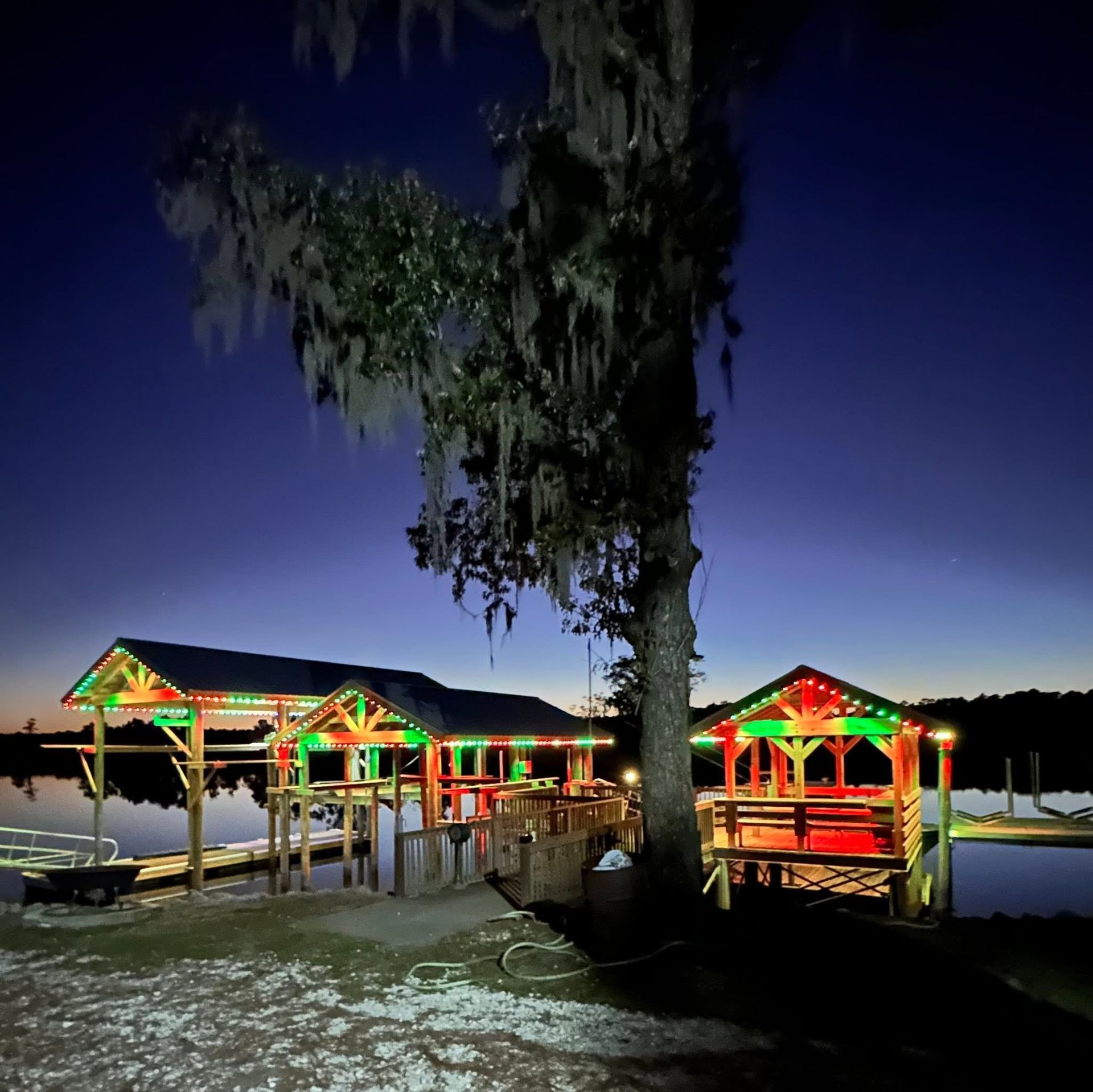Colorfully lit lakeside pavilion at dusk beside a large tree and calm water