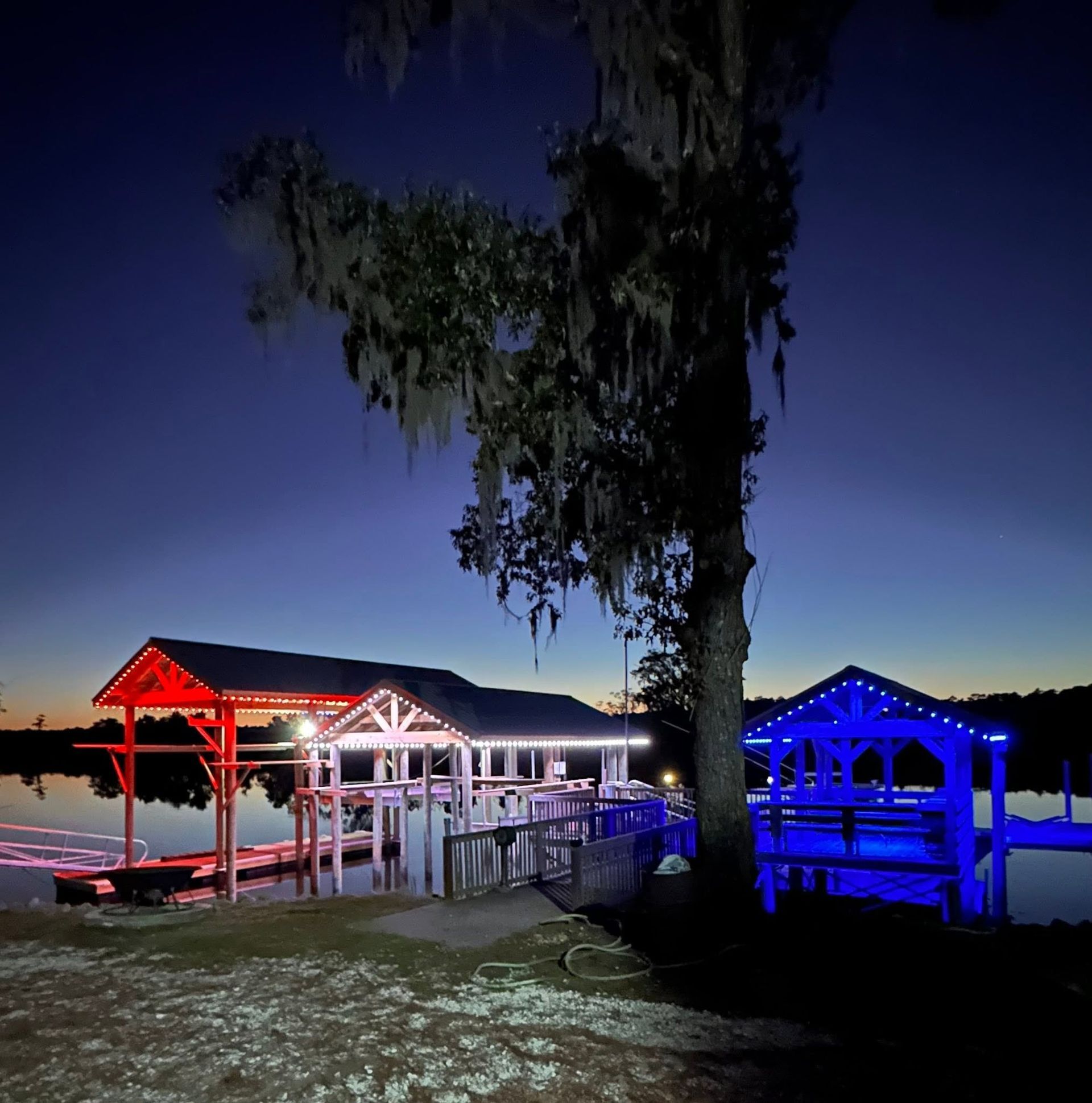 Lit dock and gazebo by the water at dusk, with red and blue lights and a tree in the foreground