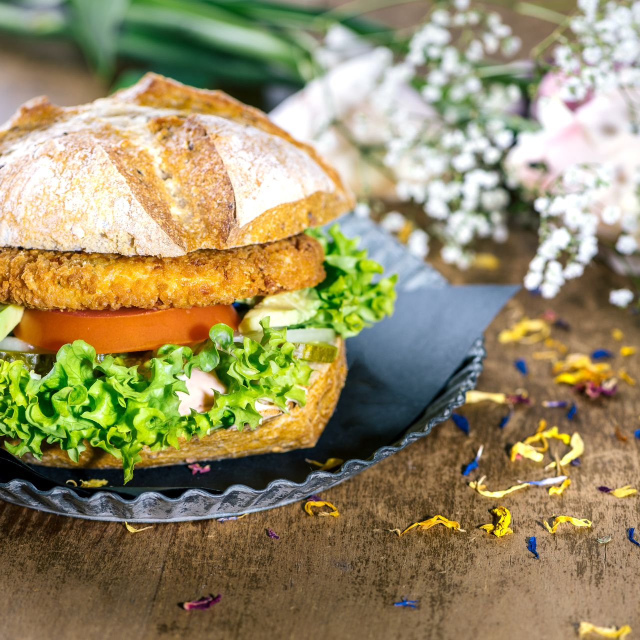 Ein paniertes Hähnchensandwich mit Salat und Tomate auf einem schwarzen Teller, angerichtet auf einem Holztisch mit floralen Akzenten.