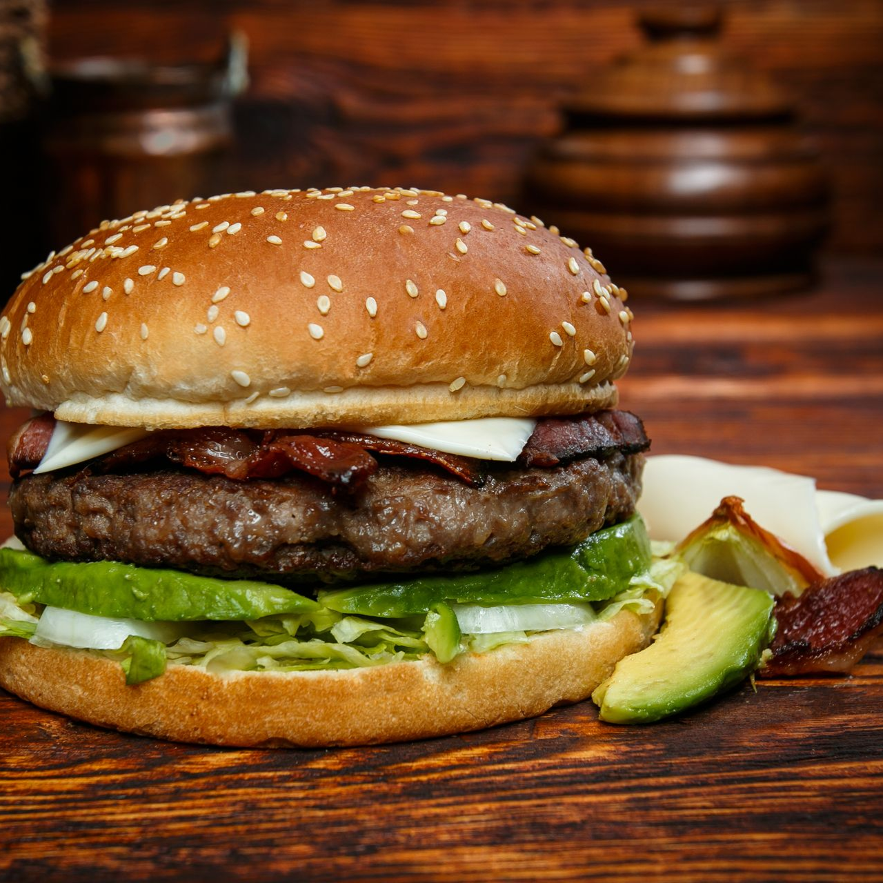 Ein Cheeseburger mit Speck und Avocadoscheiben auf einem Sesambrötchen, serviert auf einem rustikalen Holztisch.