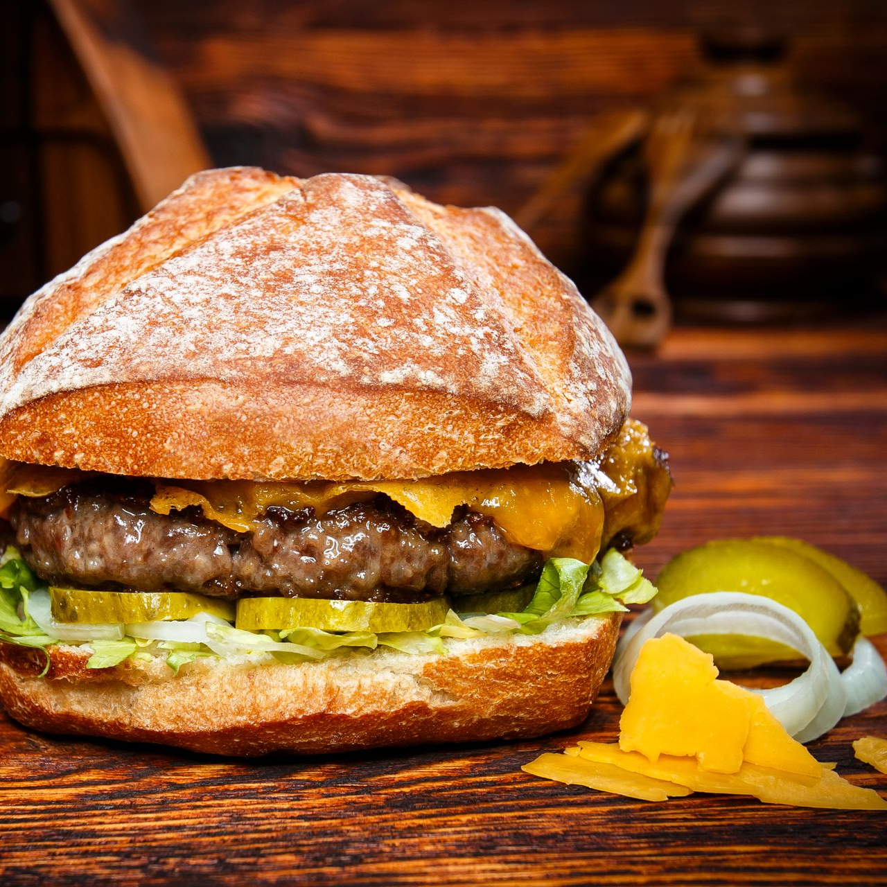 Ein Cheeseburger auf einem rustikalen Holztisch, dazu Gurkenscheiben und Zwiebelringe.