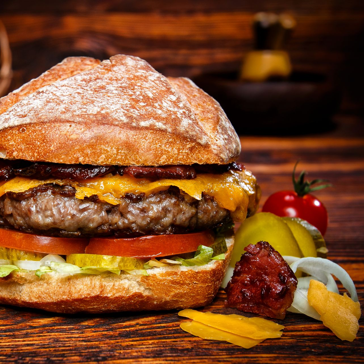 Ein Cheeseburger mit Speck, Tomaten, Salat und Essiggurken auf einem Holztisch, dazu eine Kirschtomate und Zwiebeln als Beilage.