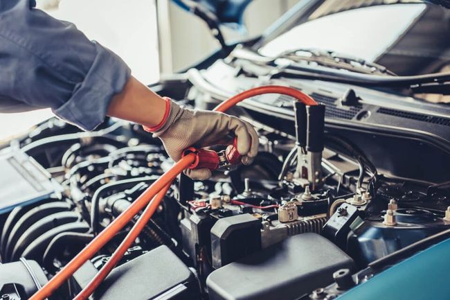 Mechanic with gloves connecting jumper cables to a car battery in an engine bay.