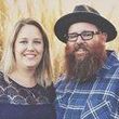 Woman with blonde hair and man with a beard smile next to each other outdoors. The man wears a hat and plaid shirt.