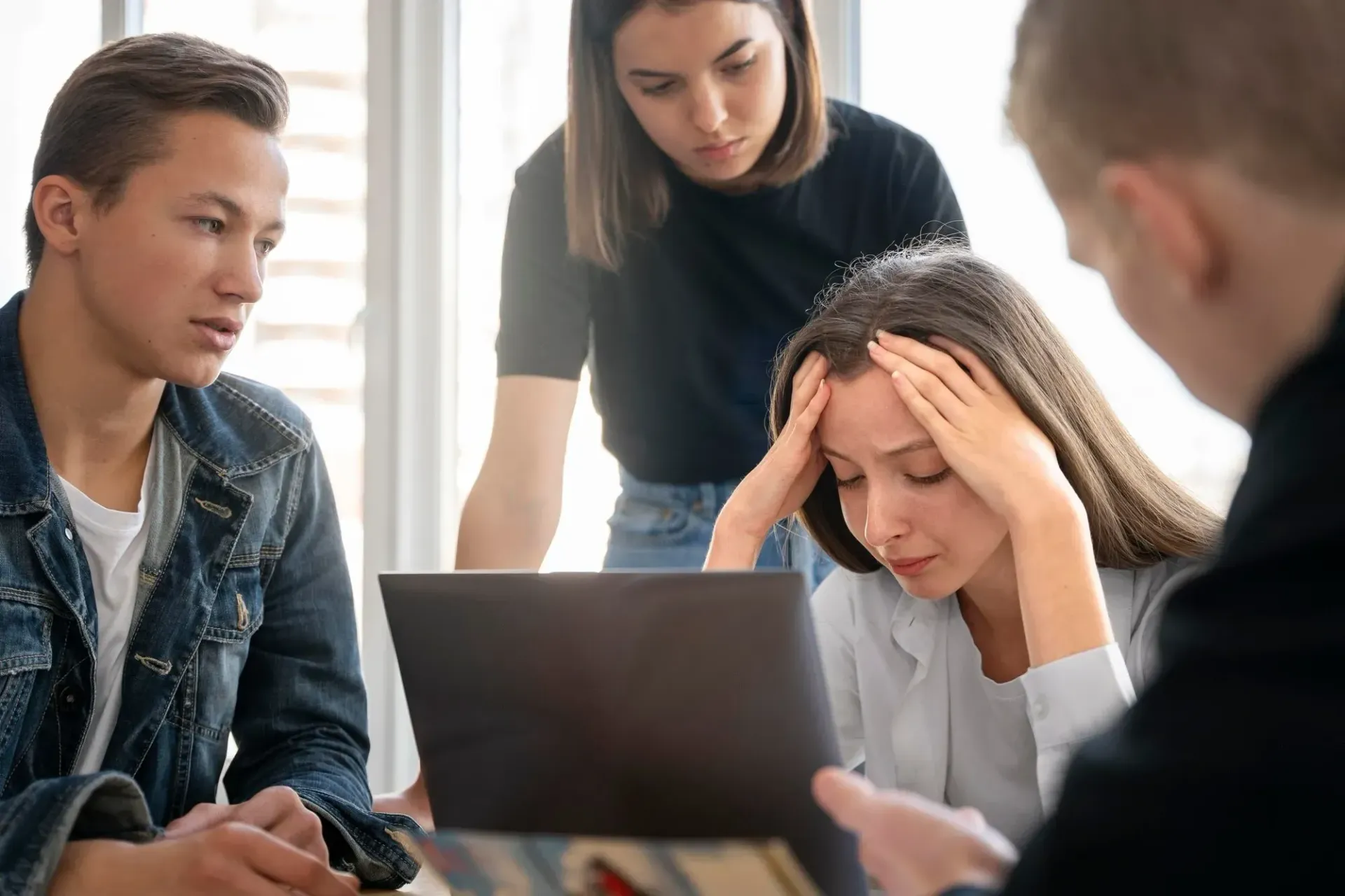 Colleagues in a meeting, one woman holding her head in stress while others look on near a laptop