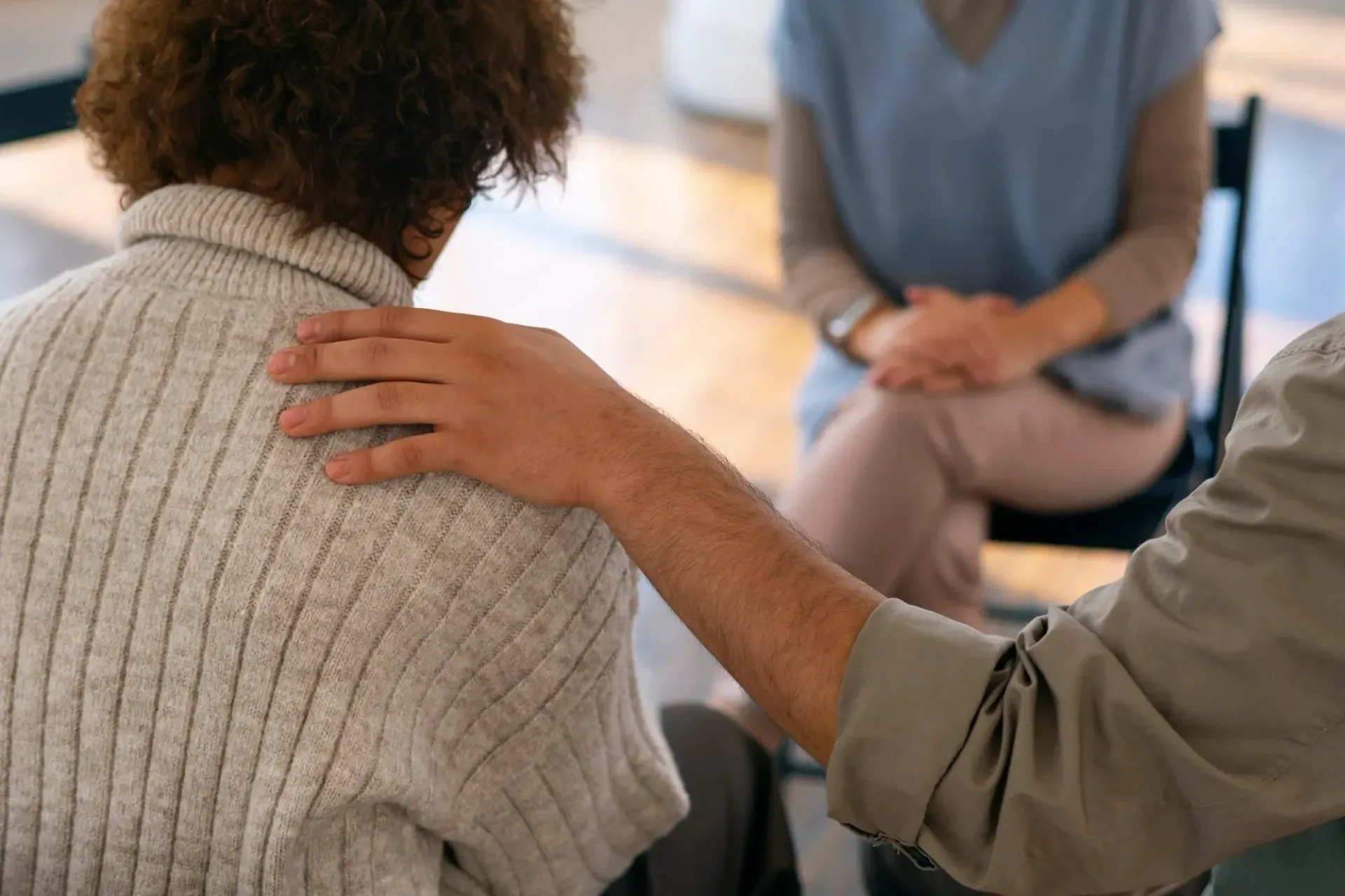 Hand on shoulder in a support group circle, with seated people in a bright room