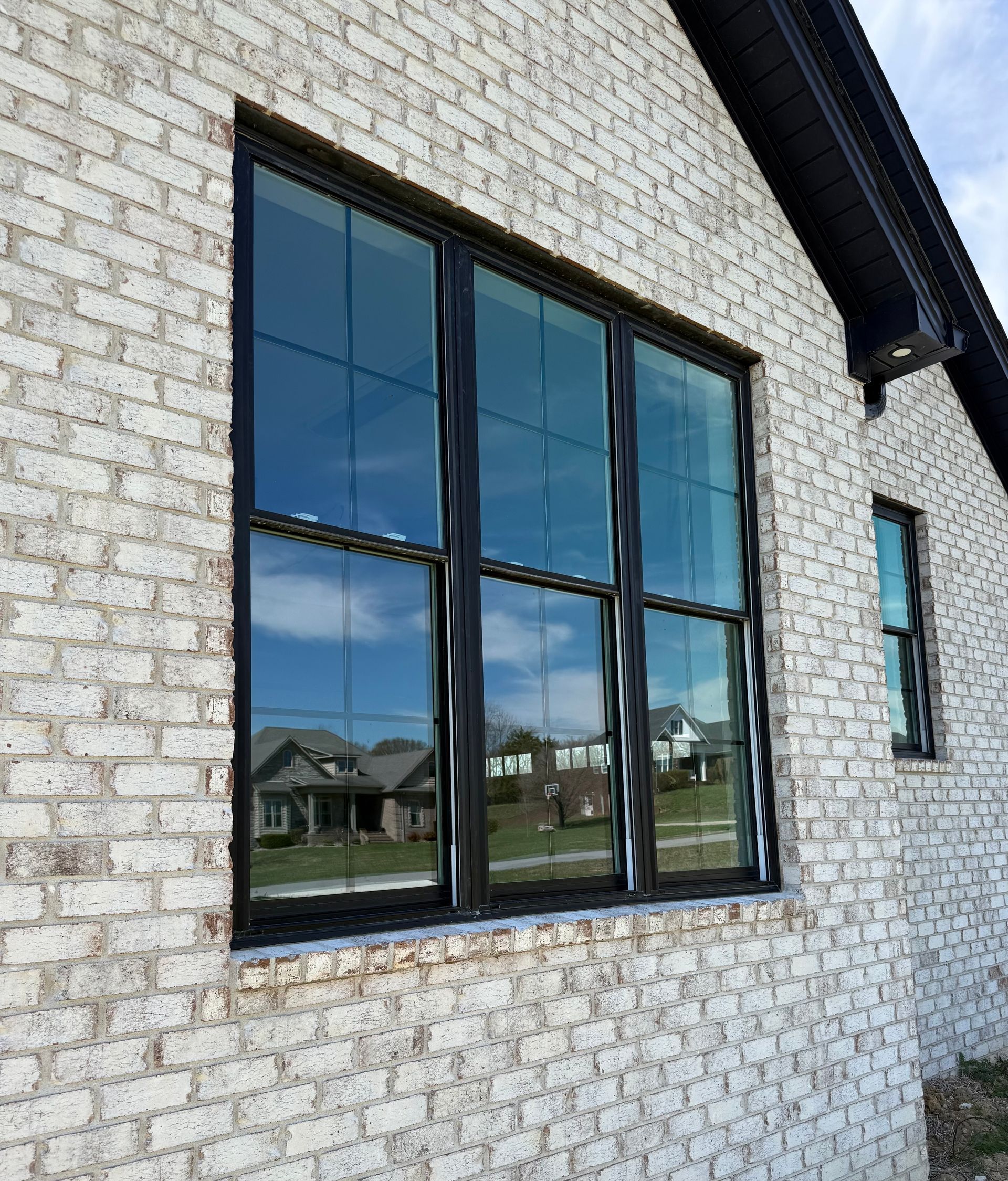 A large, three-paneled black-framed window on a wall made of textured, light-colored brick with a dark roof overhang.