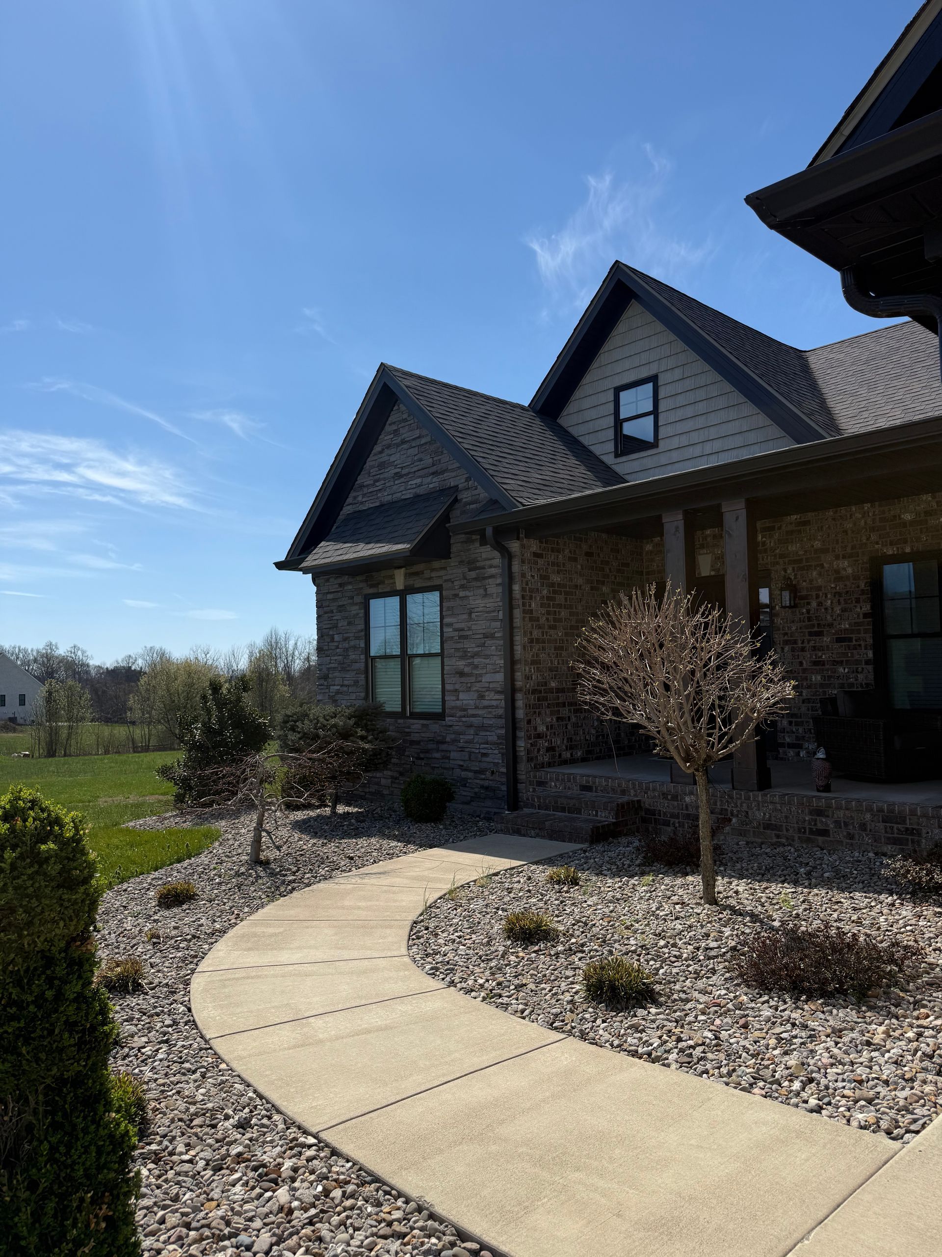 A brick home with a curved concrete walkway, gravel landscaping, and bare trees under a bright blue sky.