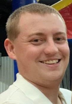 Smiling man with short blonde hair wearing a white shirt, indoors.