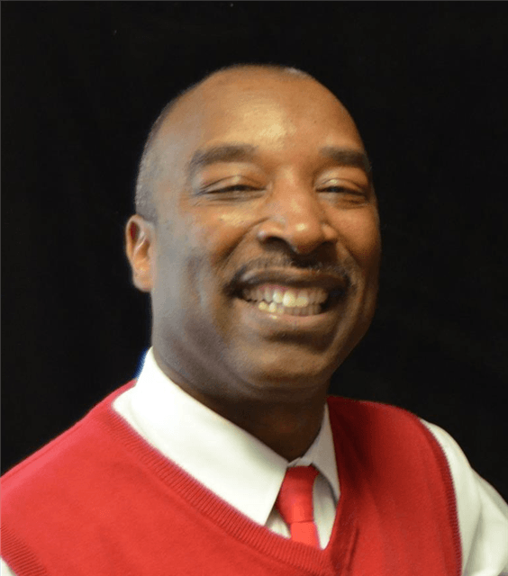 Man with dark skin smiling, wearing a red vest over a white shirt and red tie. Black background.