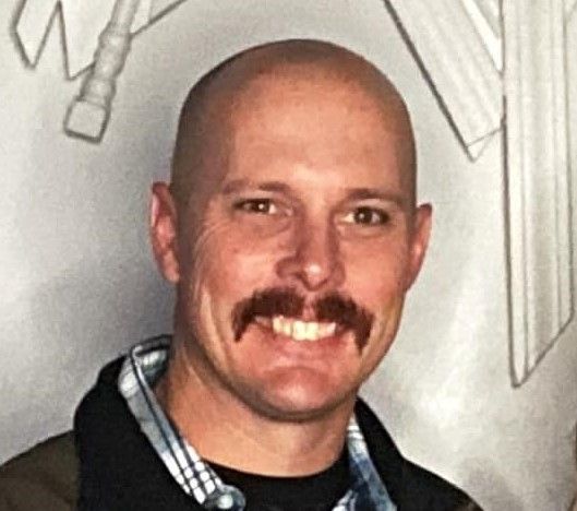 Bald man with a mustache smiles; wearing black jacket over plaid shirt.