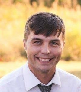 Man with short brown hair, smiling, wearing a white shirt and tie; blurred outdoor background.