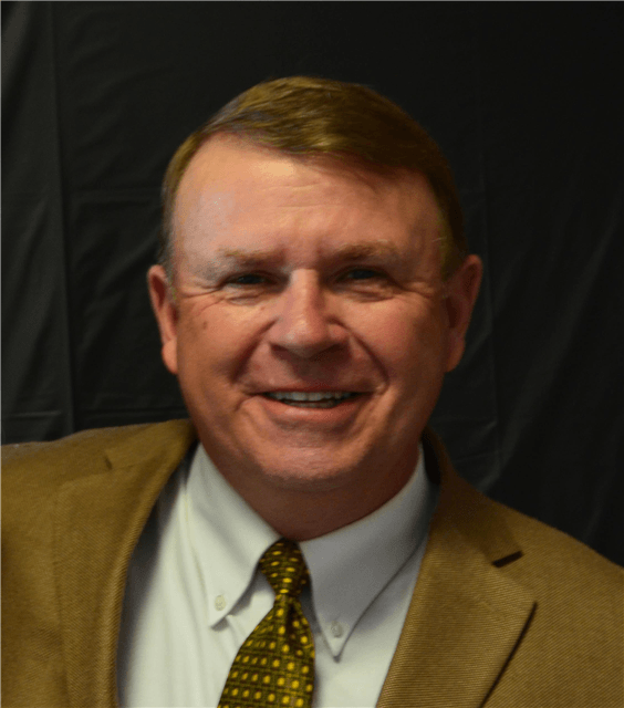 Smiling man in a tan jacket, white shirt, and patterned gold tie against a dark background.