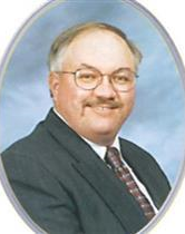 Man in suit, wearing glasses, smiling slightly, framed in an oval border.