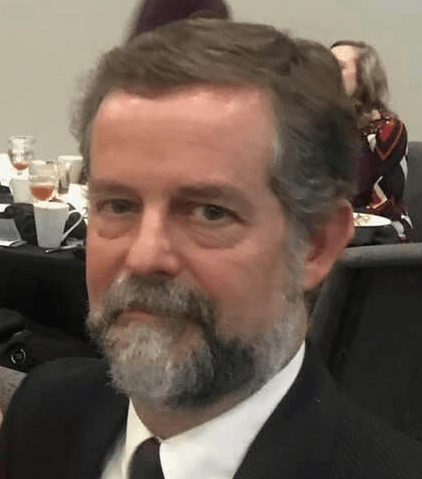 Man with graying hair and beard, wearing a suit, seated at a table in a meeting.