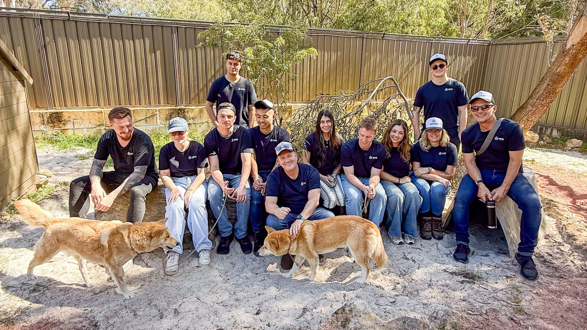 Group of people in navy shirts pose with two dogs in a sandy enclosure.