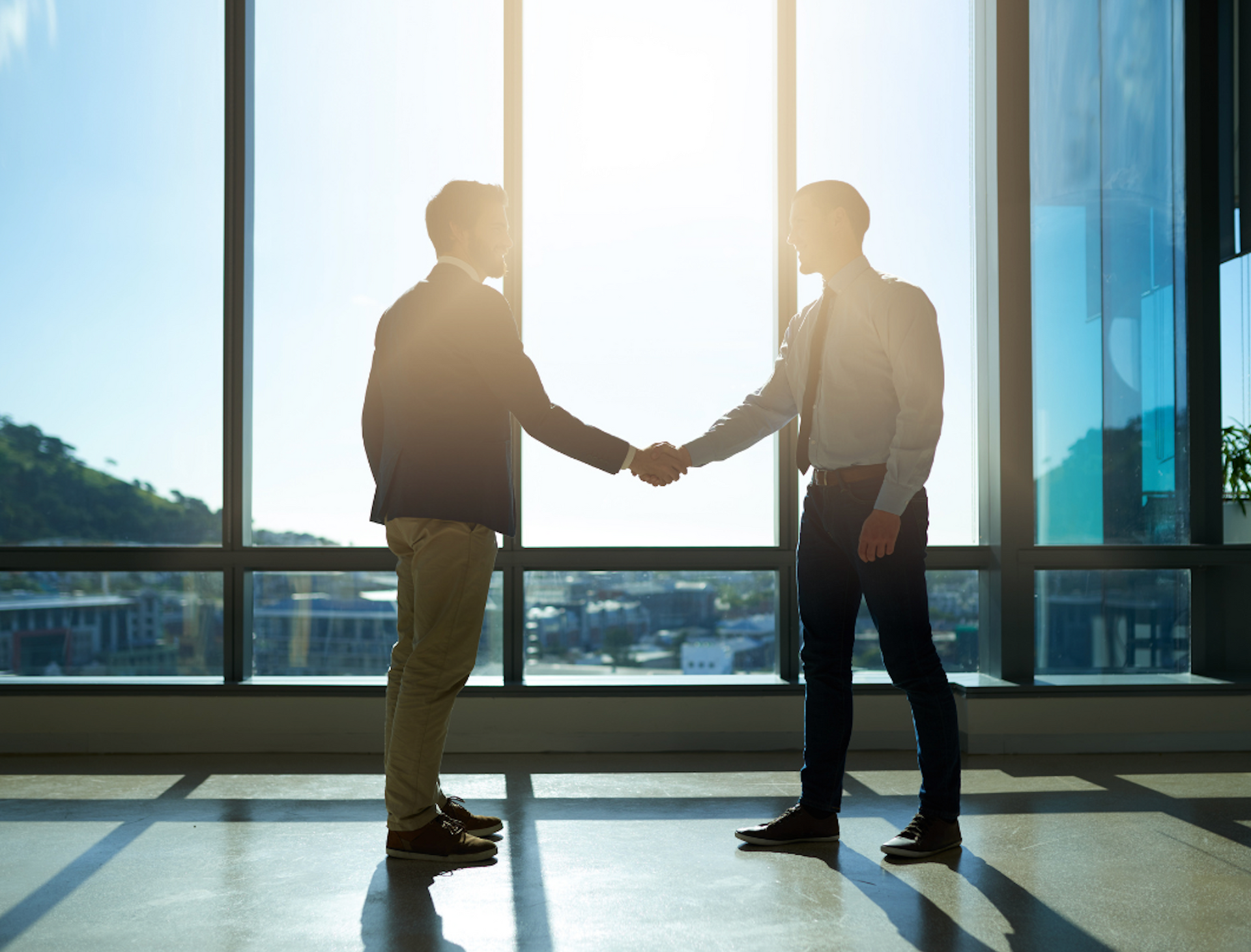 Two men are shaking hands in front of a large window.