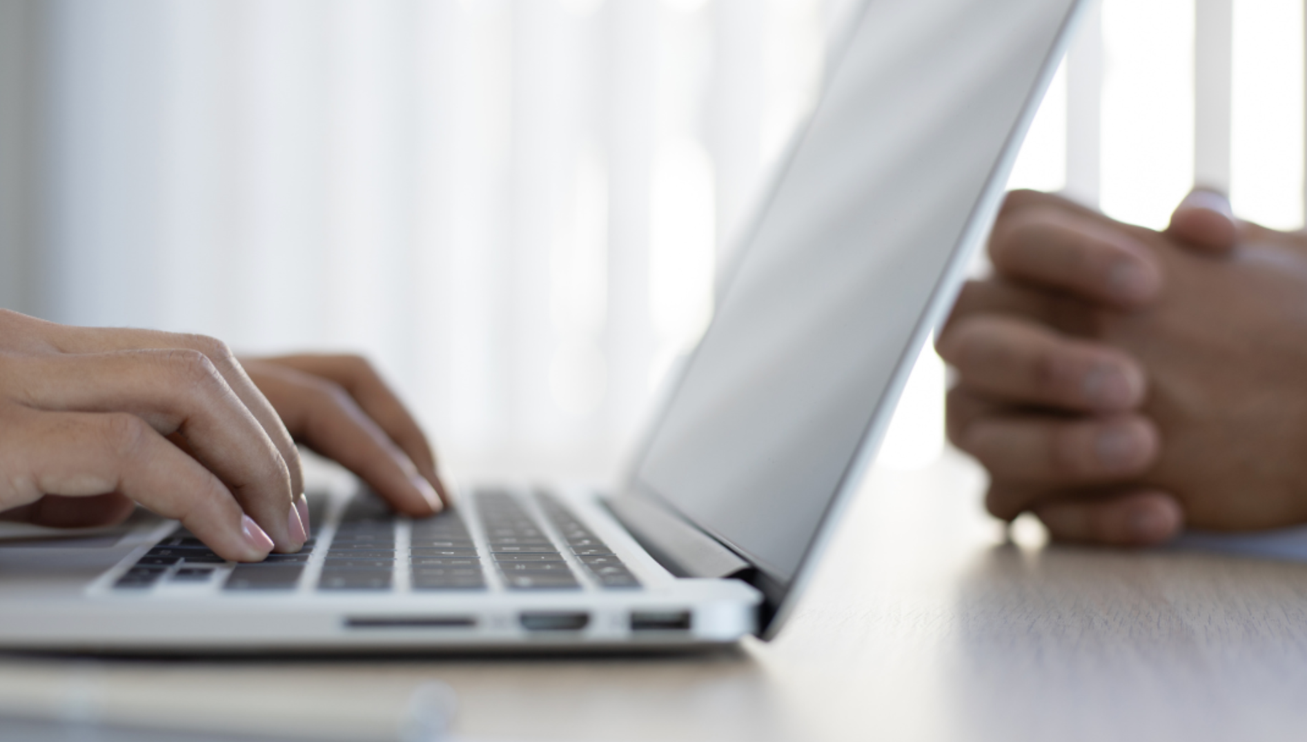 Hands typing on a laptop; another person's hands are out of focus to the side.