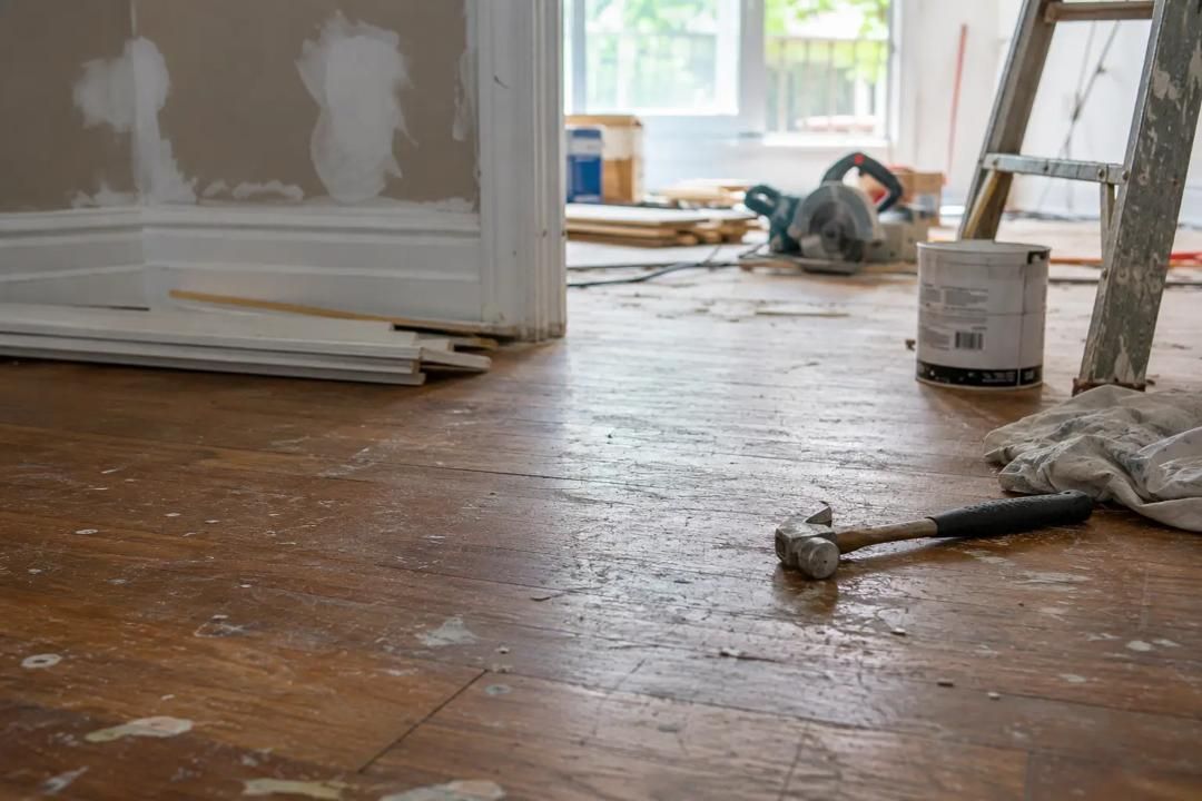 Partially renovated room with hardwood floor, drywall, paint cans, and tools near a window