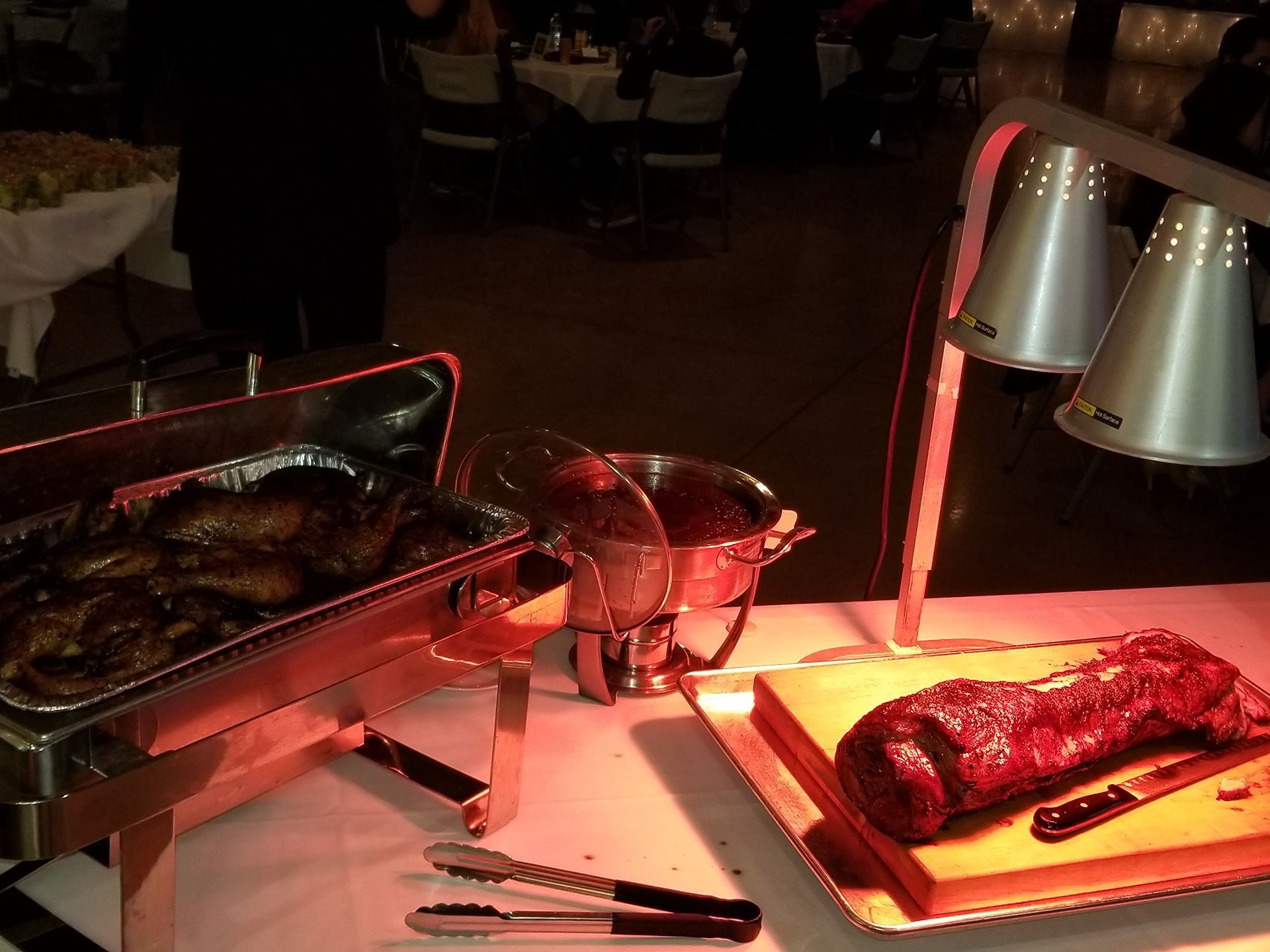 Dimly lit buffet table with grilled meat under heat lamps and serving utensils nearby