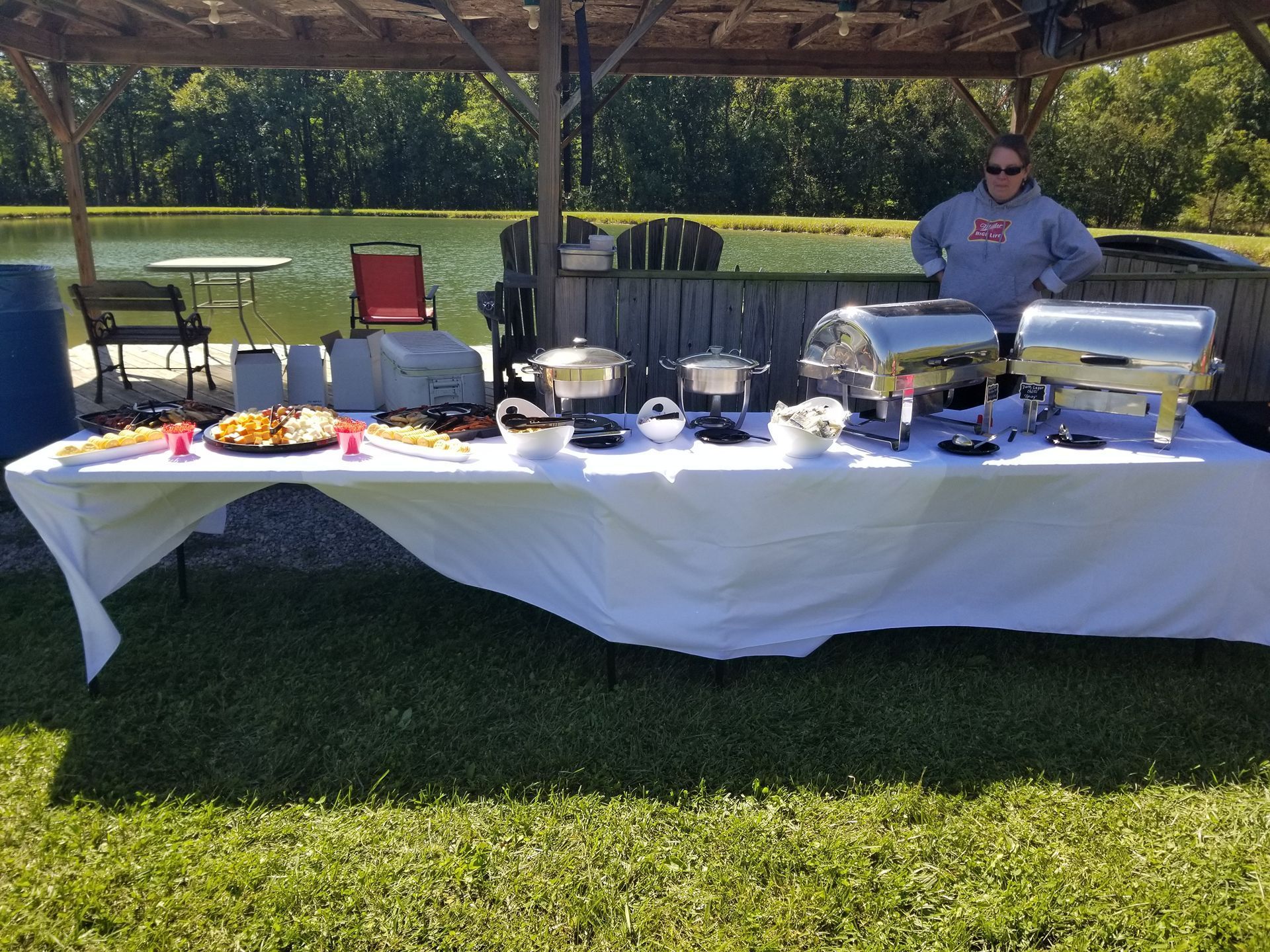 Outdoor buffet table under a pergola with covered dishes and drinks, attended by a person in a blue shirt