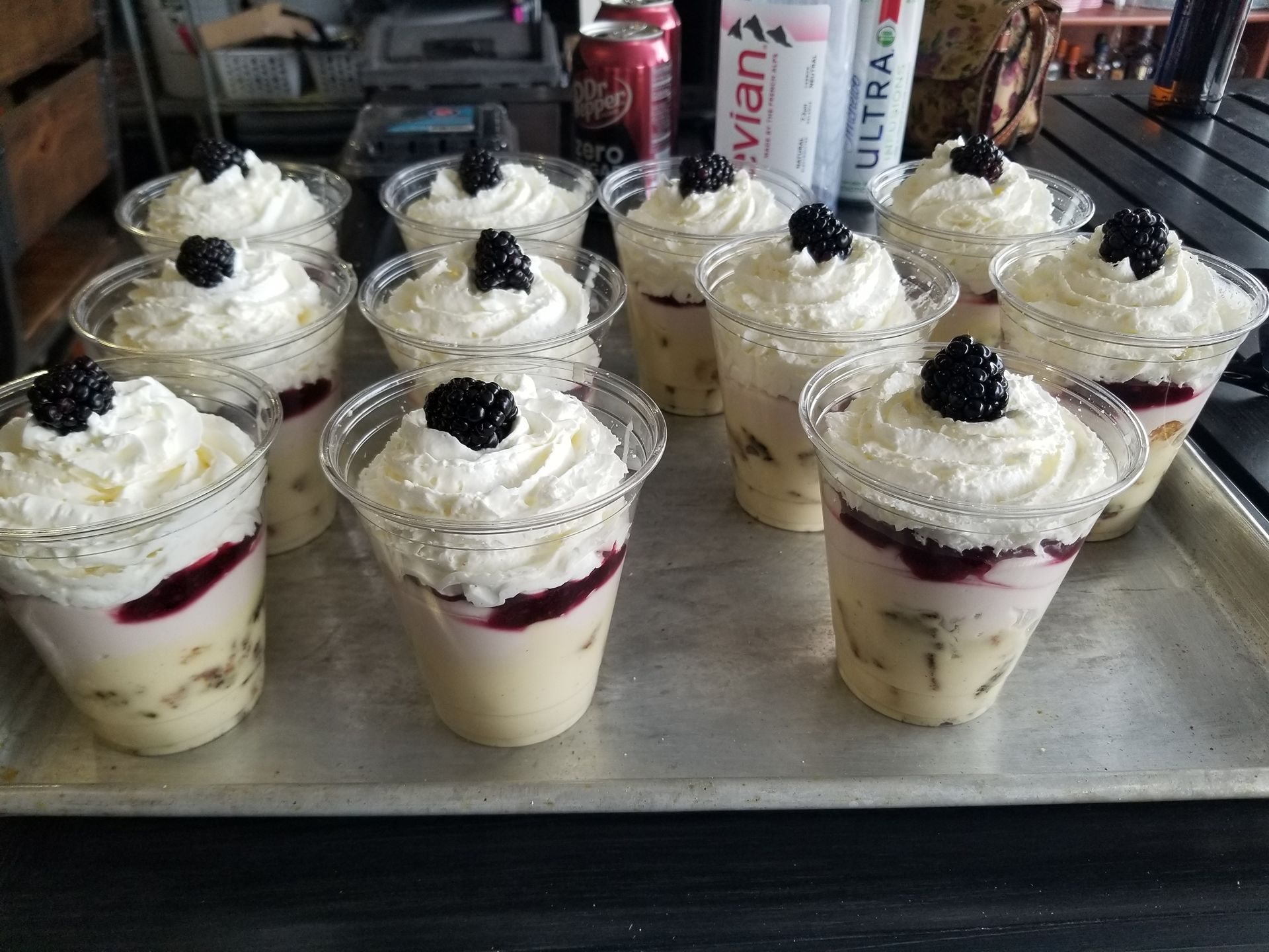 Assorted dessert cups topped with whipped cream and blackberries on a tray in a bakery display