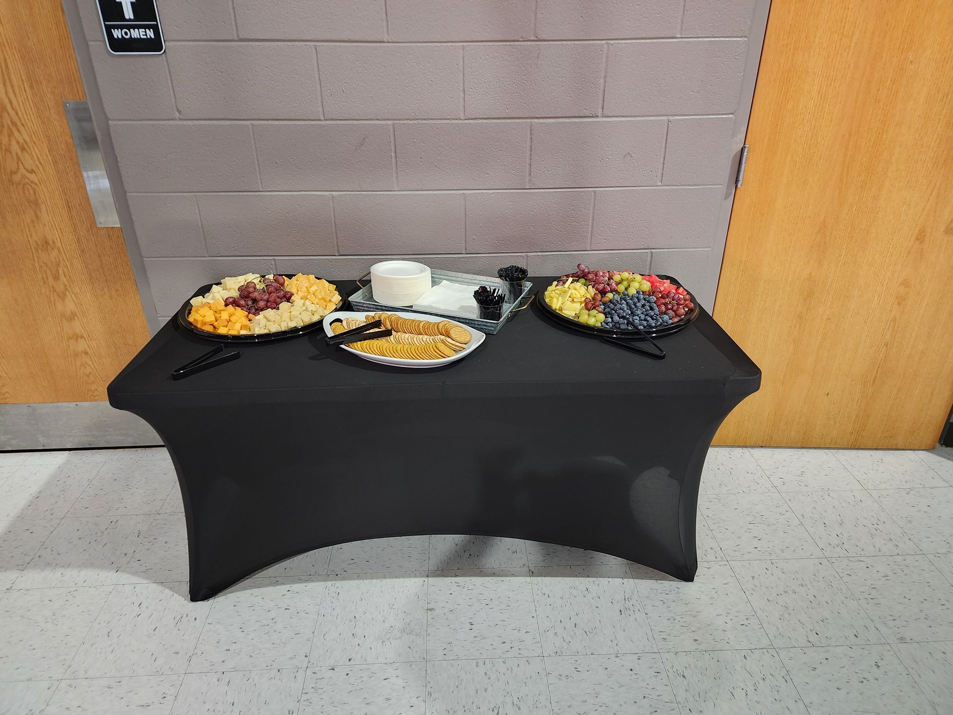 Buffet table with plates of food and drinks on a black cloth, set against a brick wall and wood doors