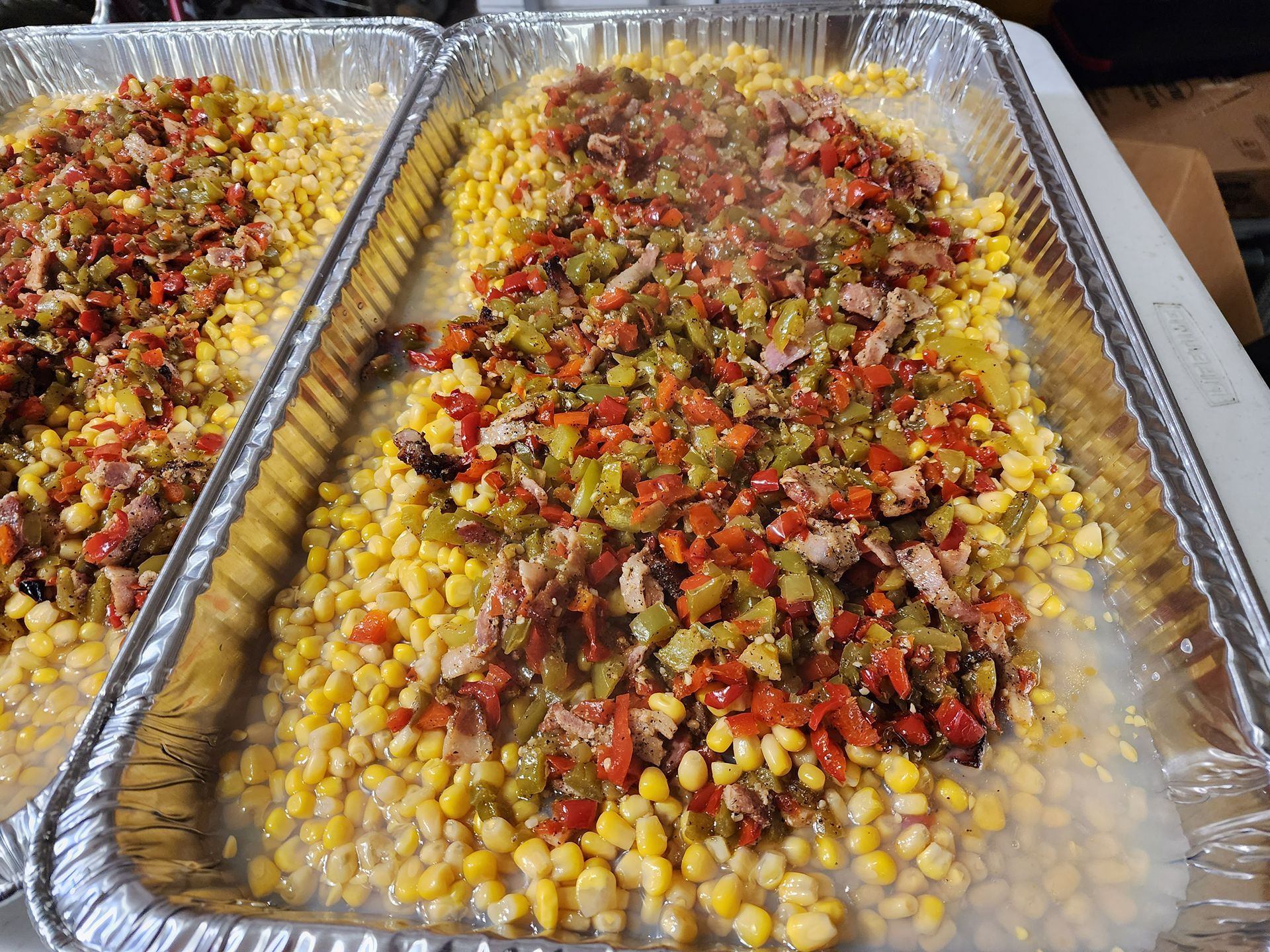Two foil trays of corn topped with diced peppers and seasonings, arranged side by side on a table