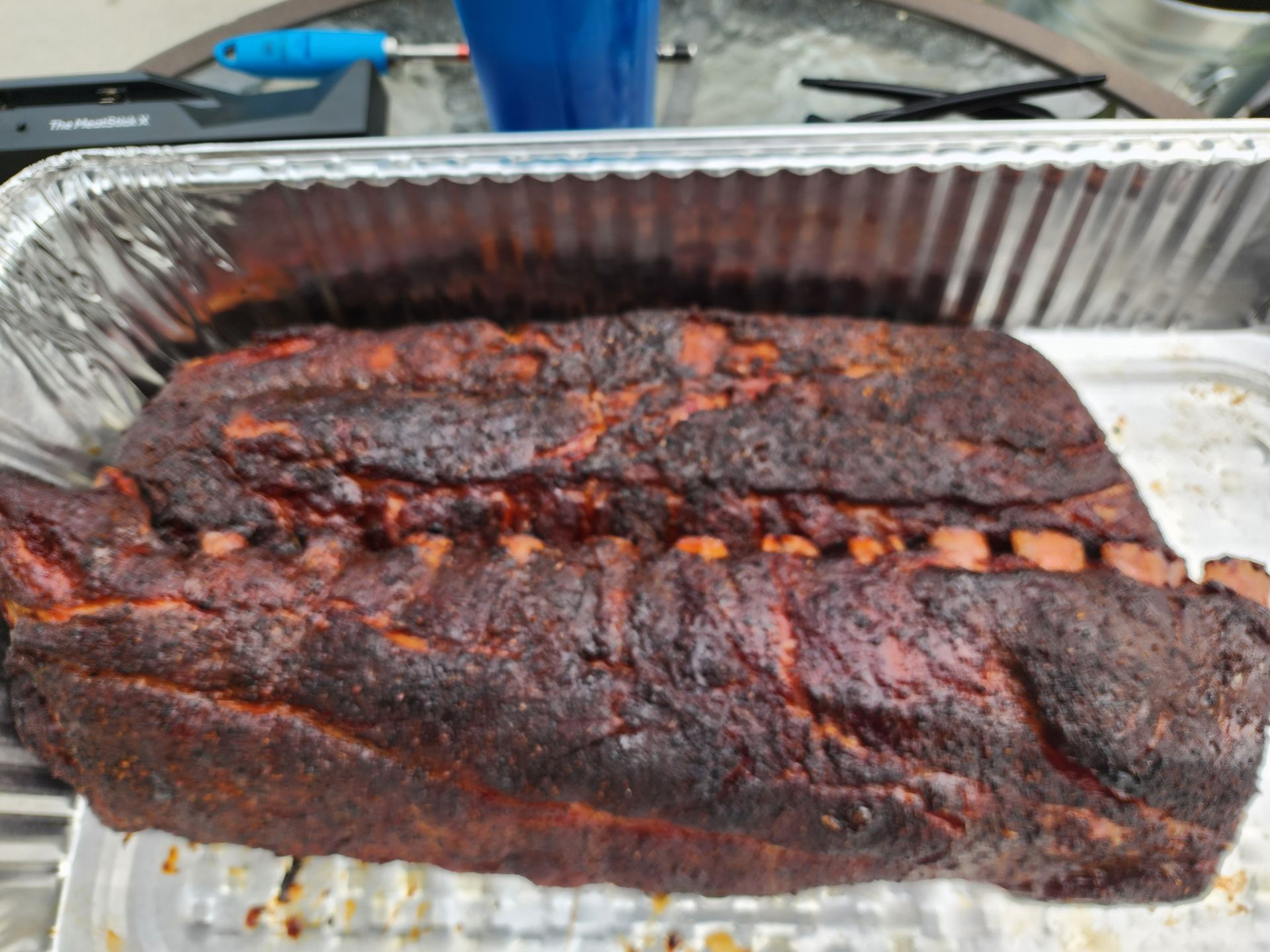 Charred smoked brisket in a foil tray on a grill, with a dark bark and a sliced seam showing meat inside