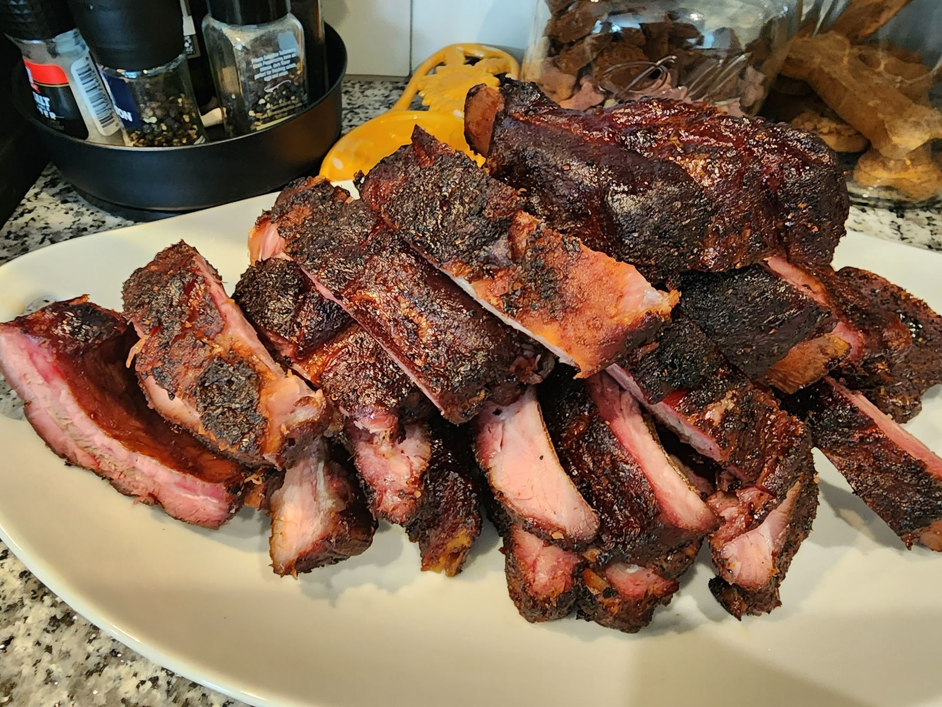 Plated sliced smoked brisket with dark bark and pink smoke ring on a white plate, barbecue spread in the background