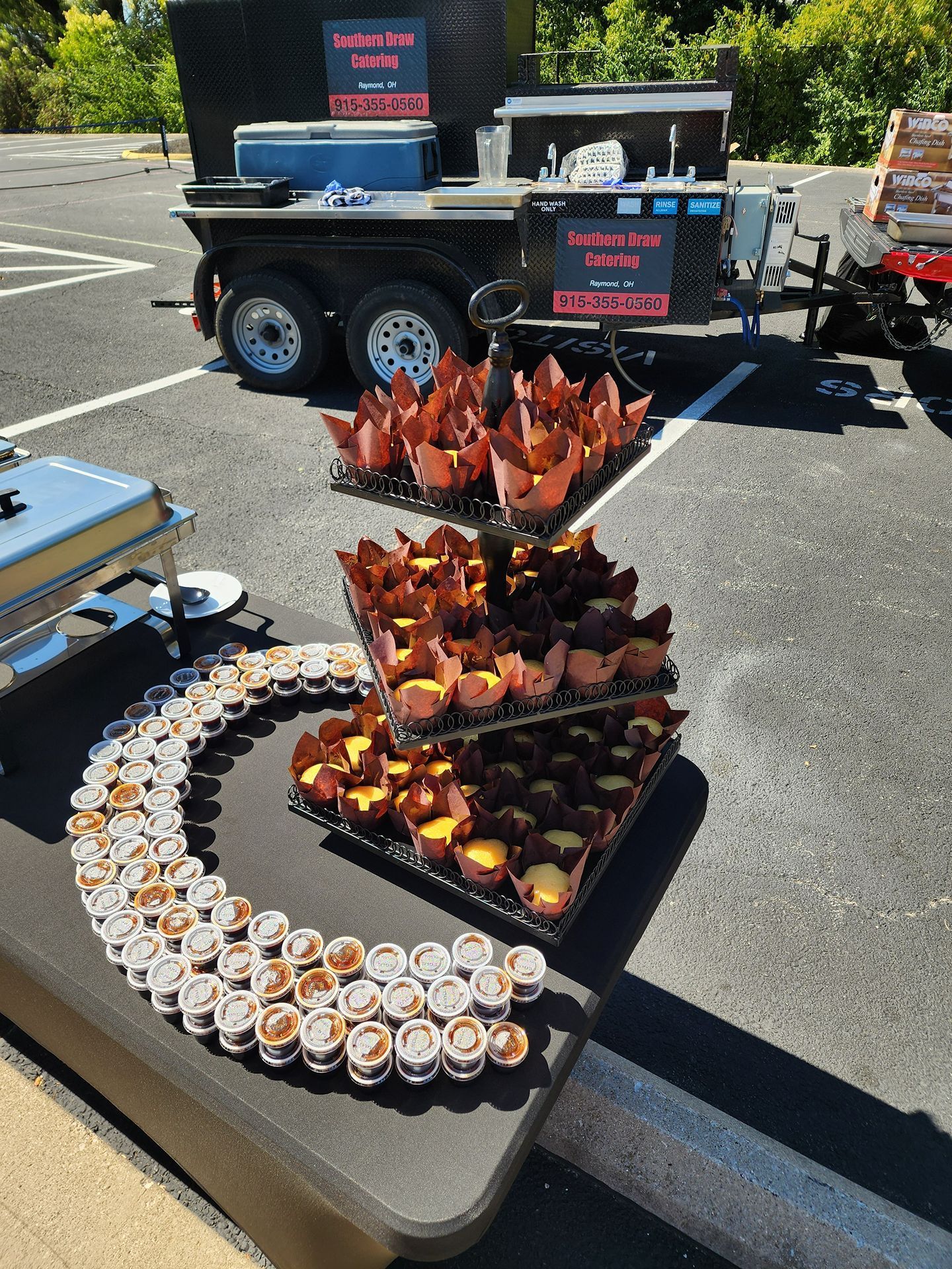 Dessert display on a table at an outdoor event, with trays of pastries and fruit cups рядом a trailer.
