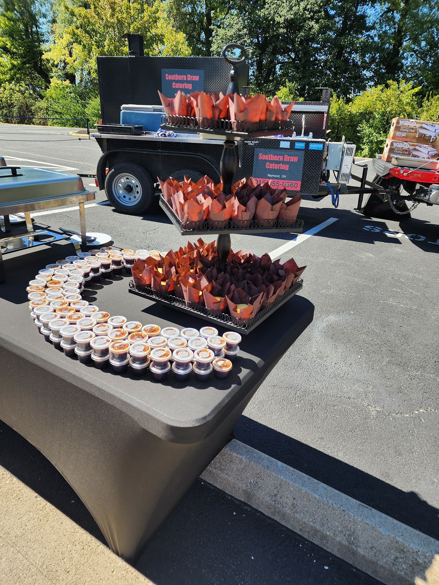 Outdoor dessert display with cupcakes and chocolate-covered treats on a table and cart in a parking lot