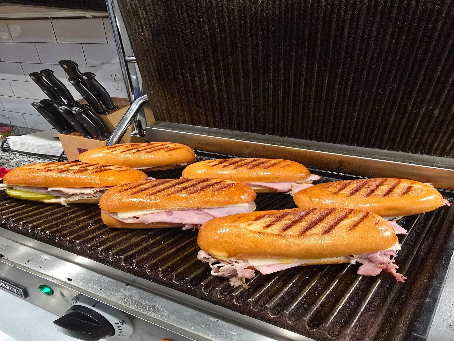 Six grilled sandwiches toasting on a commercial grill with a raised lid and skewers at the back.