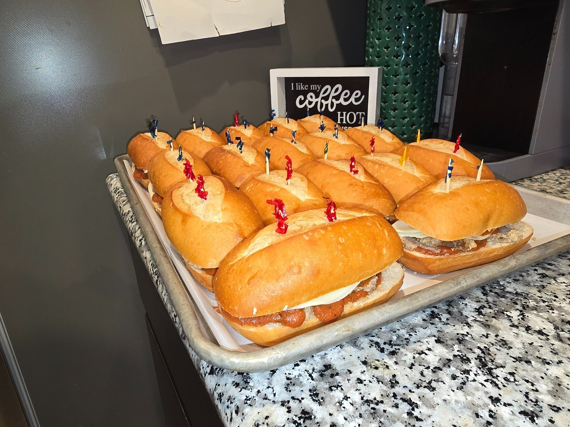 Tray of small sandwiches with red toothpicks on a granite counter, with a coffee sign in the background