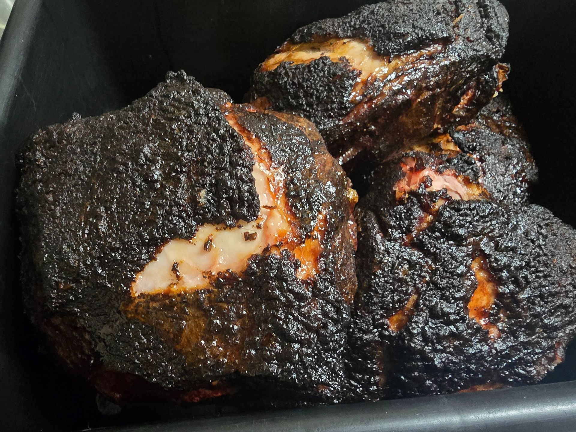 Charred barbecued meat pieces in a dark tray, with smoky black crust and bits of orange-brown interior