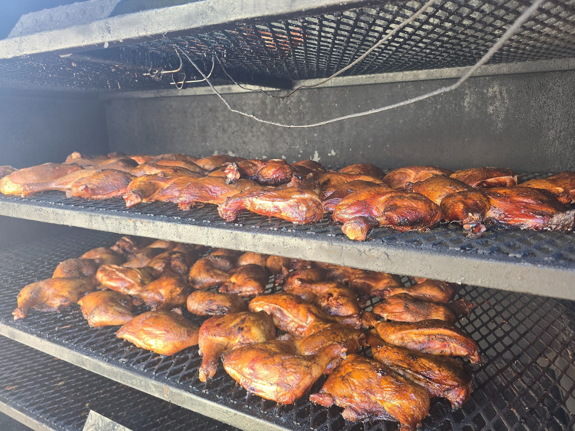 Rows of glazed chicken wings grilling on metal racks in a smoker, with visible flames and smoke.