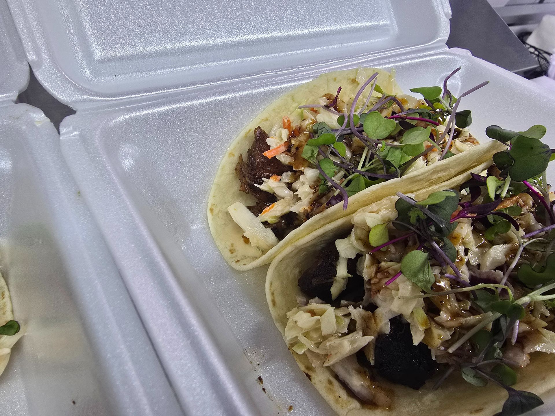 Two tacos in a white foam takeout container, topped with greens and diced onions.