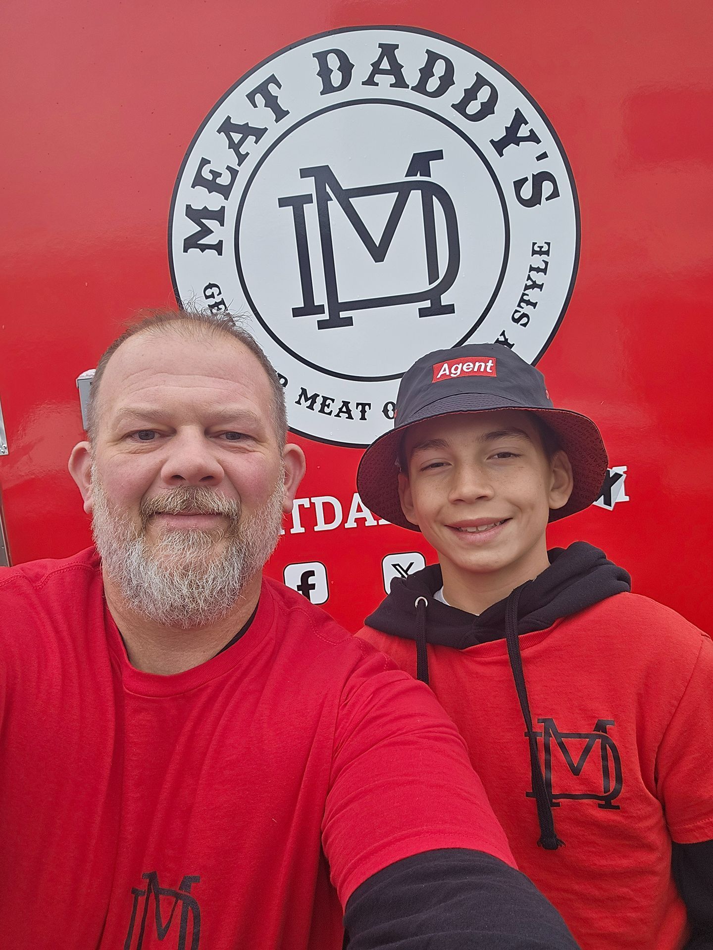 Two people in red shirts smiling in front of a Meat Daddy’s logo and red backdrop