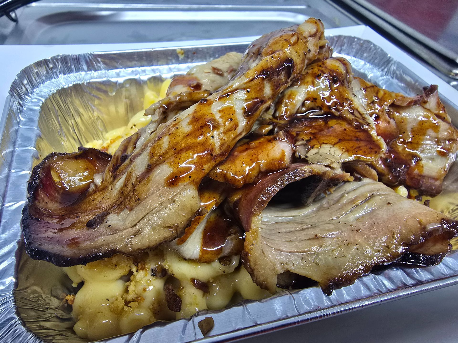 Tray of grilled meat with sauce, piled in a disposable aluminum pan on a table.