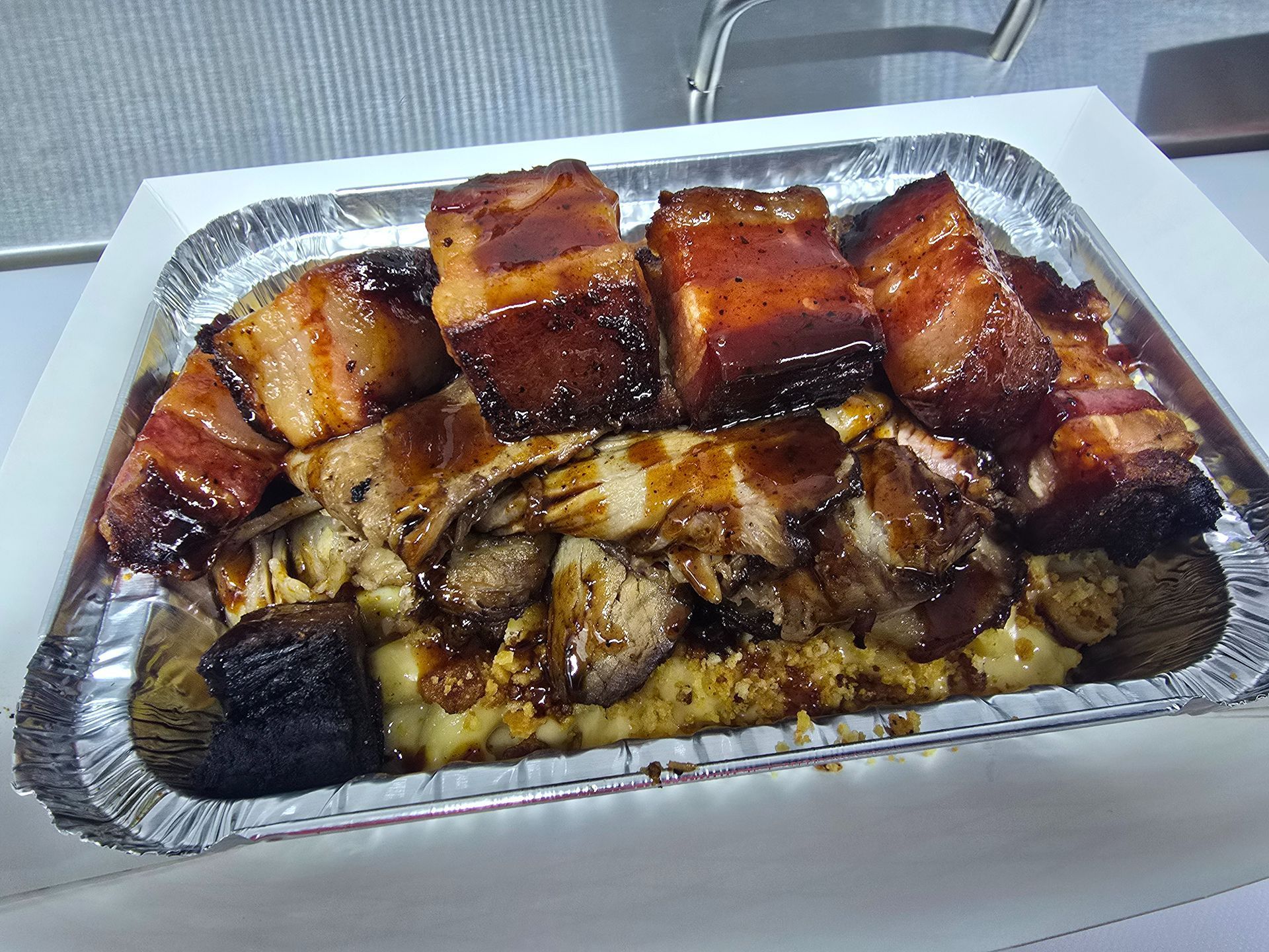 Foil tray of glazed roasted meat and charred vegetables on a white table, likely a takeout meal