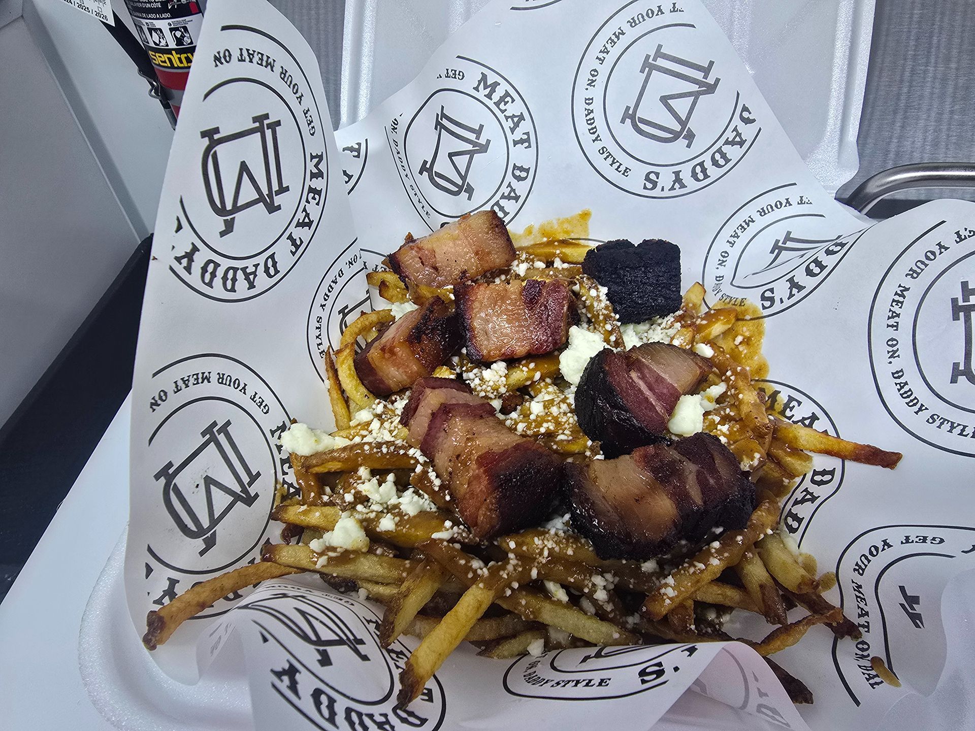Gourmet fries topped with bacon, cheese, and sauce in a branded paper tray
